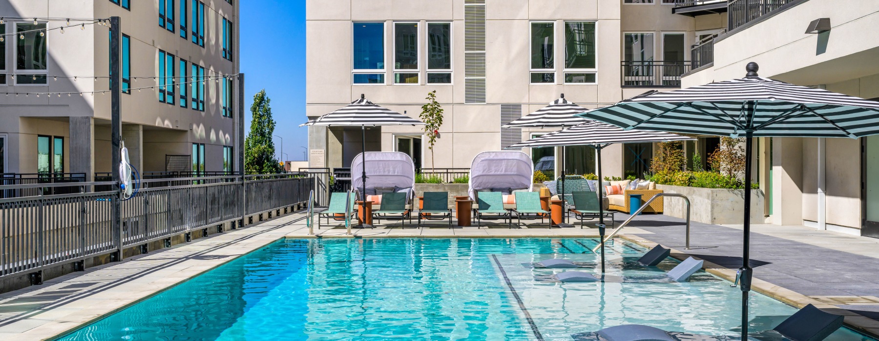 Outdoor swimming pool at Broadstone Saratoga featuring striped umbrellas, in-water lounge chairs, and modern seating areas surrounded by the apartment community.