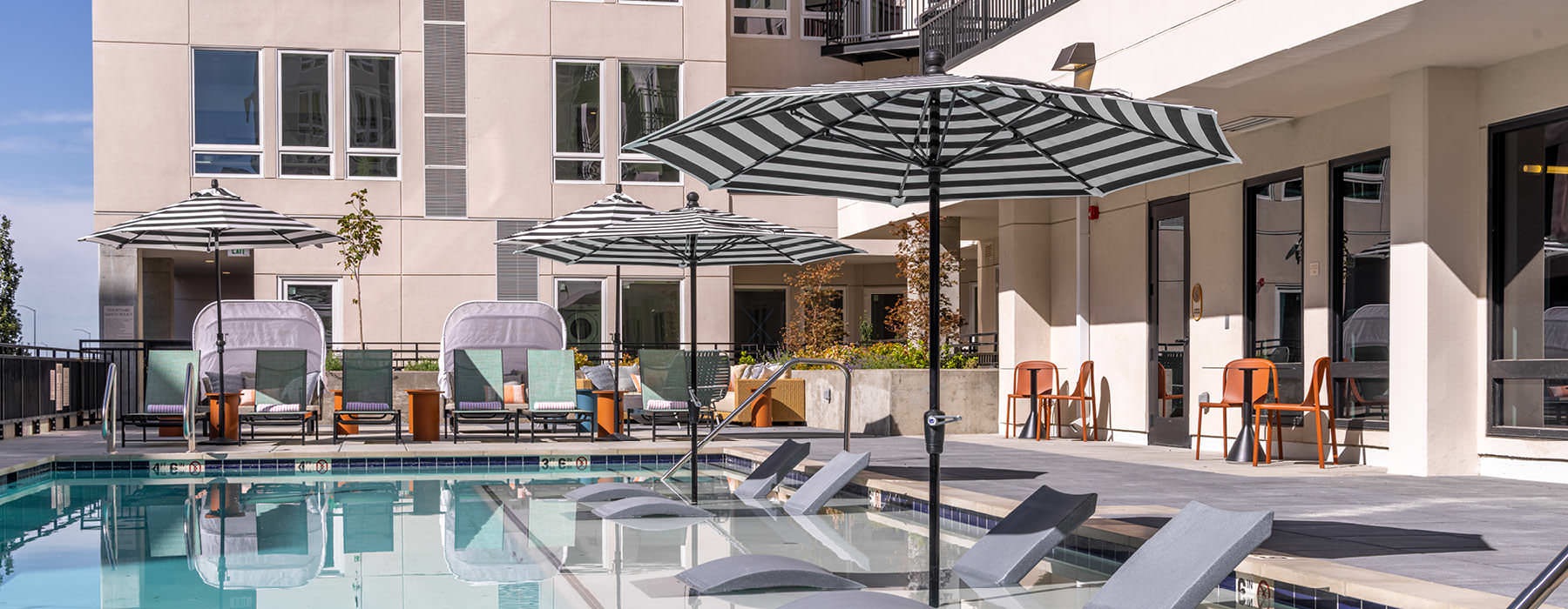 Outdoor swimming pool at Broadstone Saratoga featuring striped umbrellas, in-water lounge chairs, and modern seating areas surrounded by the apartment community.